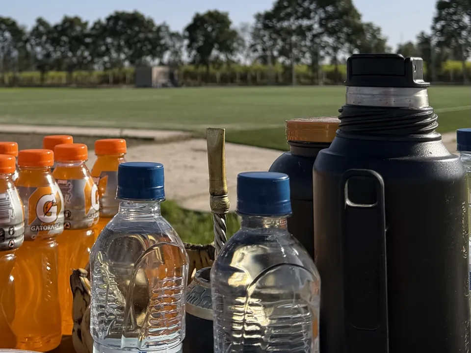 Mesa de hidratación junto a la cancha durante un entrenamiento, con botellas de agua, bebidas, un mate y un termo